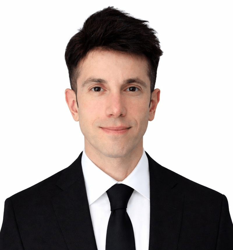 Professional headshot of Damian Rezaee wearing a black suit and tie against a light background.